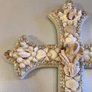 May include: A white wooden cross decorated with a mosaic of seashells. The cross is made of various sizes and shapes of shells, creating a textured and intricate design.