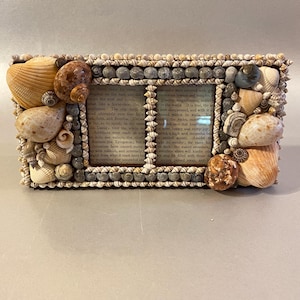 May include: A rectangular picture frame decorated with various natural seashells in shades of tan, brown, and grey. The frame features two glass openings displaying printed text from a book. The shells are arranged along the edges and center.