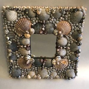 May include: A square mirror frame completely covered in a dense mosaic of various seashells in shades of gray, tan, and white. The design features larger fan-shaped scallop shells arranged in a grid pattern, surrounded by smaller round shells.