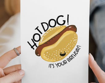 May include: A white greeting card featuring a cartoon illustration of a brown hot dog in a yellow bun with a small smiling face. The text reads HOT DOG! IT'S YOUR BIRTHDAY! in black lettering arched around the central food graphic.