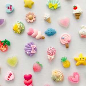 Peut inclure: Une collection d'embellissements en r&eacute;sine color&eacute;e de diff&eacute;rentes formes, notamment des c&oelig;urs, des &eacute;toiles, des coquillages, des cornets de glace et des sucettes. Les embellissements sont dans les tons de rose, jaune, bleu et blanc, parfaits pour les projets de bricolage.
