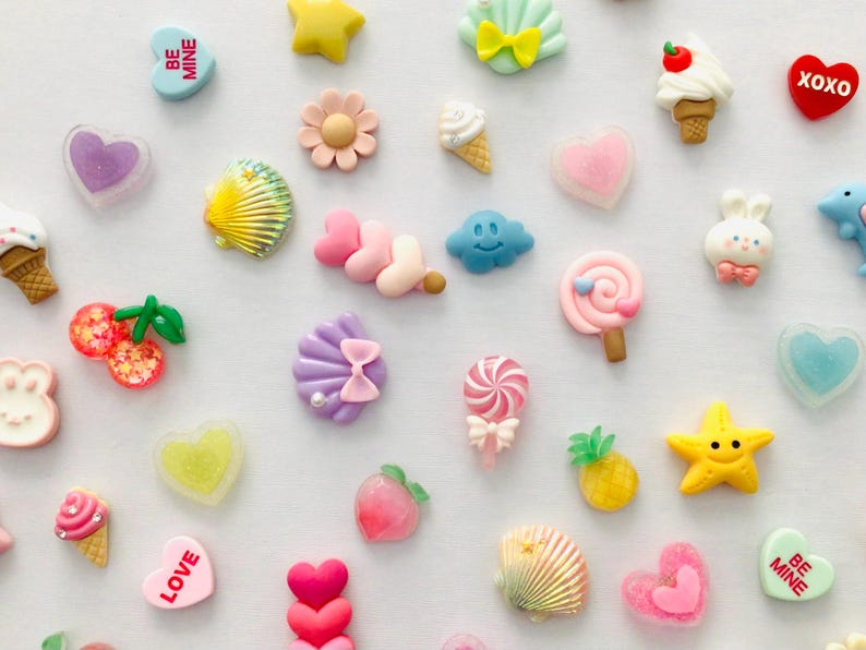 Peut inclure: Une collection d'embellissements en r&eacute;sine color&eacute;e de diff&eacute;rentes formes, notamment des c&oelig;urs, des &eacute;toiles, des coquillages, des cornets de glace et des sucettes. Les embellissements sont dans les tons de rose, jaune, bleu et blanc, parfaits pour les projets de bricolage.