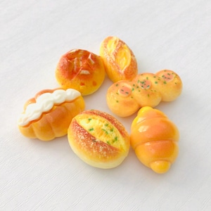 May include: A collection of miniature bread items, including a croissant, rolls, and loaves, in shades of yellow and orange. Some pieces have white or green accents, arranged on a white surface.