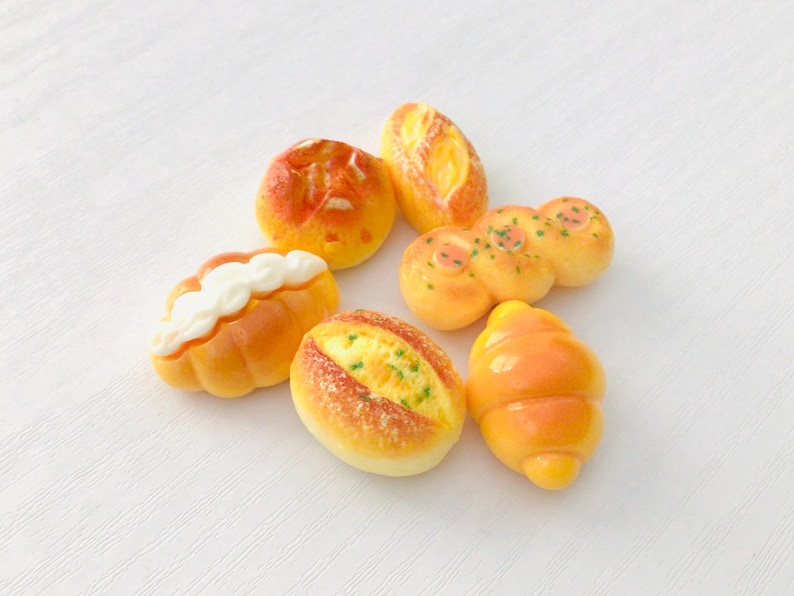May include: A collection of miniature bread items, including a croissant, rolls, and loaves, in shades of yellow and orange. Some pieces have white or green accents, arranged on a white surface.
