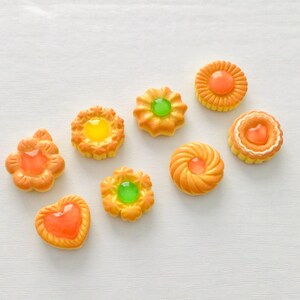 May include: Overhead view of eight miniature, decorative biscuits in orange and yellow hues. They feature various shapes and clear, jelly-like centres in orange, yellow, and green. The biscuits are arranged on a white surface.