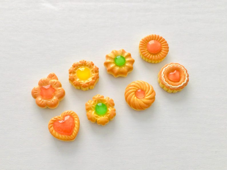 May include: Overhead view of eight miniature, decorative biscuits in orange and yellow hues. They feature various shapes and clear, jelly-like centres in orange, yellow, and green. The biscuits are arranged on a white surface.
