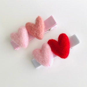 May include: Three felt heart hair clips in pink, light pink, and red. The clips are attached to pink and white ribbon.