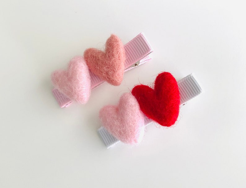May include: Three felt heart hair clips in pink, light pink, and red. The clips are attached to pink and white ribbon.