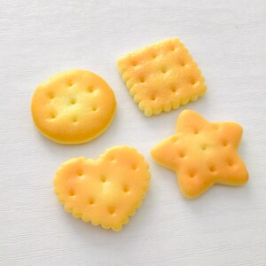May include: Four golden-yellow crackers in various shapes: a circle, a square, a heart, and a star. Each biscuit has small holes and a slightly textured surface. The biscuits are arranged on a white surface.