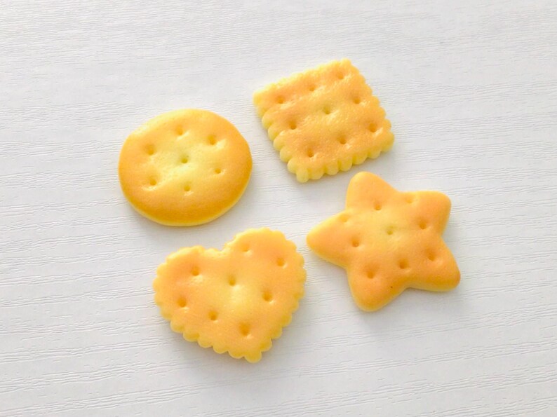 May include: Four golden-yellow crackers in various shapes: a circle, a square, a heart, and a star. Each biscuit has small holes and a slightly textured surface. The biscuits are arranged on a white surface.