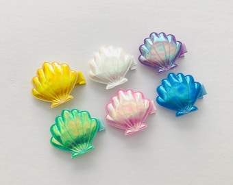 May include: Six colourful seashell hair clips in yellow, white, pink, green, purple, and blue. Each clip has a shimmering, iridescent finish. The clips are arranged on a white surface.