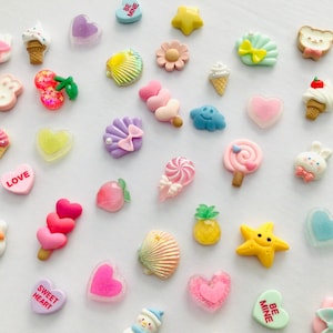 Peut inclure: Une collection de breloques en r&eacute;sine color&eacute;es de diff&eacute;rentes formes, notamment des c&oelig;urs, des cornets de glace, des coquillages et des &eacute;toiles. Certaines breloques comportent des textes comme "LOVE" et "XOXO". Les breloques sont dans des tons de rose, jaune, bleu et blanc.