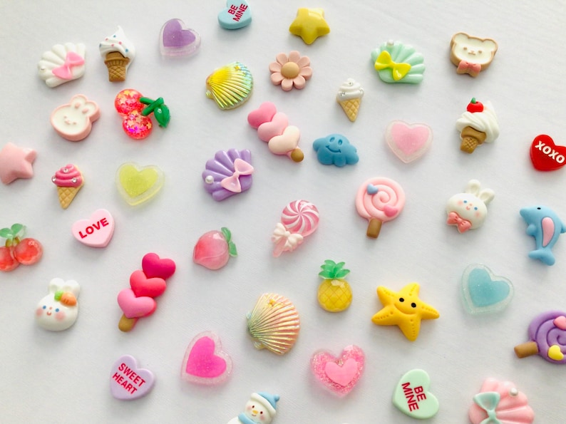 Peut inclure: Une collection de breloques en r&eacute;sine color&eacute;es de diff&eacute;rentes formes, notamment des c&oelig;urs, des cornets de glace, des coquillages et des &eacute;toiles. Certaines breloques comportent des textes comme "LOVE" et "XOXO". Les breloques sont dans des tons de rose, jaune, bleu et blanc.