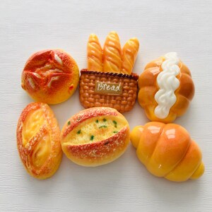 May include: A collection of miniature bread items, including a round bun, a baguette basket with the word "Bread", a roll with white icing, a loaf, a bread roll with green specks, and a croissant. The bread items are orange and yellow.