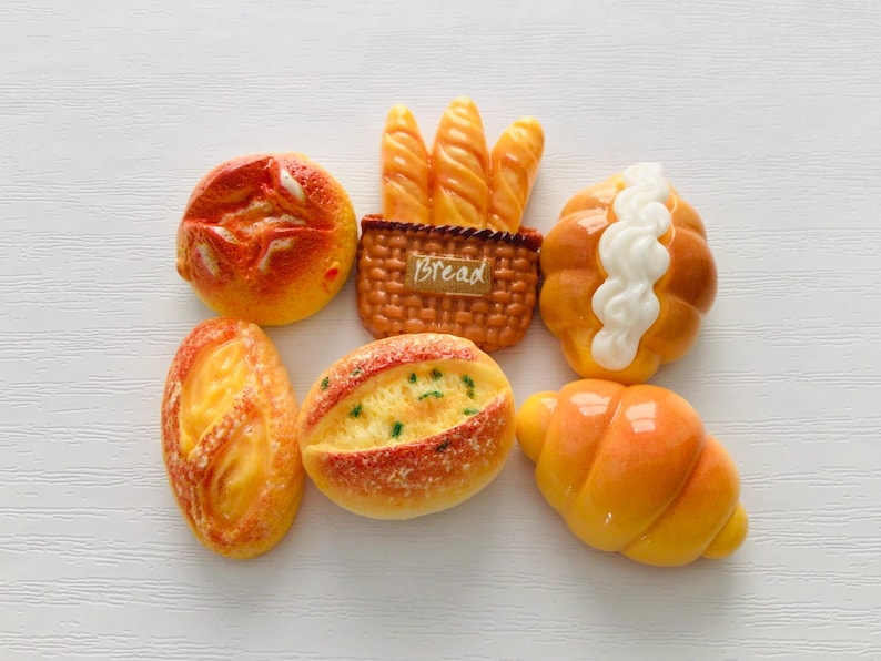 May include: A collection of miniature bread items, including a round bun, a baguette basket with the word "Bread", a roll with white icing, a loaf, a bread roll with green specks, and a croissant. The bread items are orange and yellow.