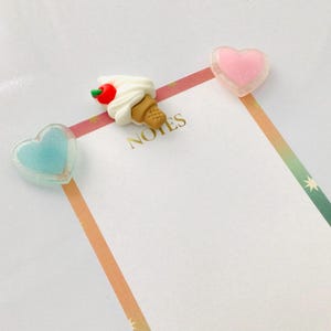 Peut inclure: Un carnet avec des trombones en forme de c&oelig;ur roses et bleus, et un trombone en forme de cornet de glace. Le mot "NOTES" est imprim&eacute; en or. Une bordure ray&eacute;e color&eacute;e ajoute une touche d&eacute;corative.