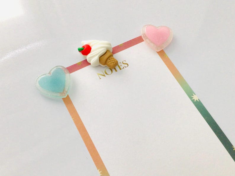 Peut inclure: Un carnet avec des trombones en forme de c&oelig;ur roses et bleus, et un trombone en forme de cornet de glace. Le mot "NOTES" est imprim&eacute; en or. Une bordure ray&eacute;e color&eacute;e ajoute une touche d&eacute;corative.