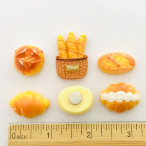 May include: A collection of miniature bread-themed magnets. The set includes a croissant, a loaf, a round bread, a baguette basket with the word "Bread", and two other baked goods. The ruler shows measurements in inches.