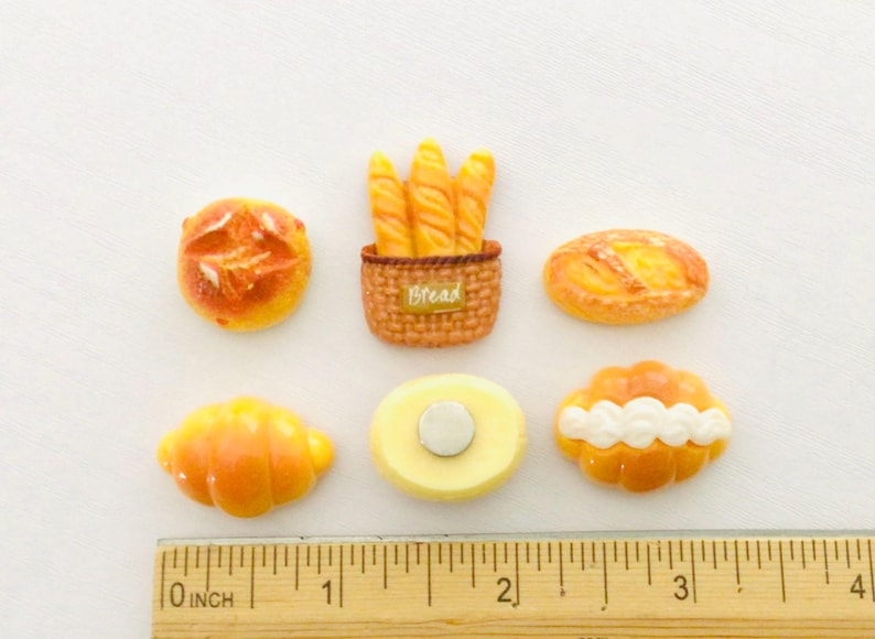 May include: A collection of miniature bread-themed magnets. The set includes a croissant, a loaf, a round bread, a baguette basket with the word "Bread", and two other baked goods. The ruler shows measurements in inches.