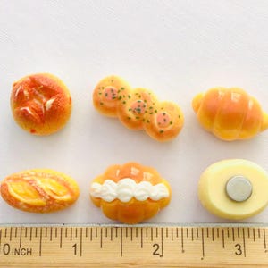 May include: A selection of miniature food magnets, including a round bun, a cluster of rolls, a croissant, a baguette, a pastry with white frosting, and a round magnet. The ruler shows the items are approximately 7.6 cm in length.