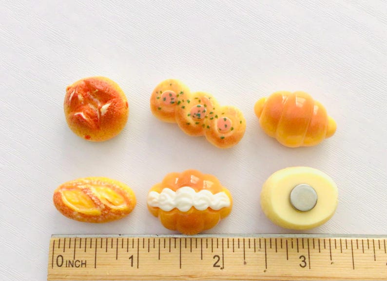 May include: A selection of miniature food magnets, including a round bun, a cluster of rolls, a croissant, a baguette, a pastry with white frosting, and a round magnet. The ruler shows the items are approximately 7.6 cm in length.