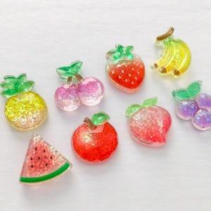 May include: Assortment of colorful, glittery fruit-shaped resin charms. Includes a pineapple, cherries, strawberry, bananas, grapes, apple, peach, and watermelon slice. Each charm has a translucent appearance with glitter accents.