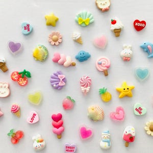 Peut inclure: Une collection d'embellissements en r&eacute;sine color&eacute;e de diff&eacute;rentes formes, notamment des c&oelig;urs, des &eacute;toiles, des cornets de glace et des coquillages. Certaines pi&egrave;ces comportent du texte tel que "BE MINE" et "XOXO". L'assortiment pr&eacute;sente des couleurs pastel et des motifs fantaisistes.