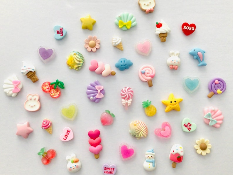 Peut inclure: Une collection d'embellissements en r&eacute;sine color&eacute;e de diff&eacute;rentes formes, notamment des c&oelig;urs, des &eacute;toiles, des cornets de glace et des coquillages. Certaines pi&egrave;ces comportent du texte tel que "BE MINE" et "XOXO". L'assortiment pr&eacute;sente des couleurs pastel et des motifs fantaisistes.