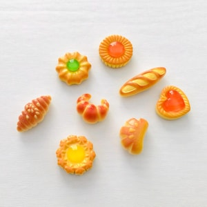 May include: A selection of miniature baked goods, including croissants, bread, and tarts, in shades of orange and yellow. The pastries are arranged on a white surface, showcasing their detailed designs and textures. A delightful assortment of miniature food items.