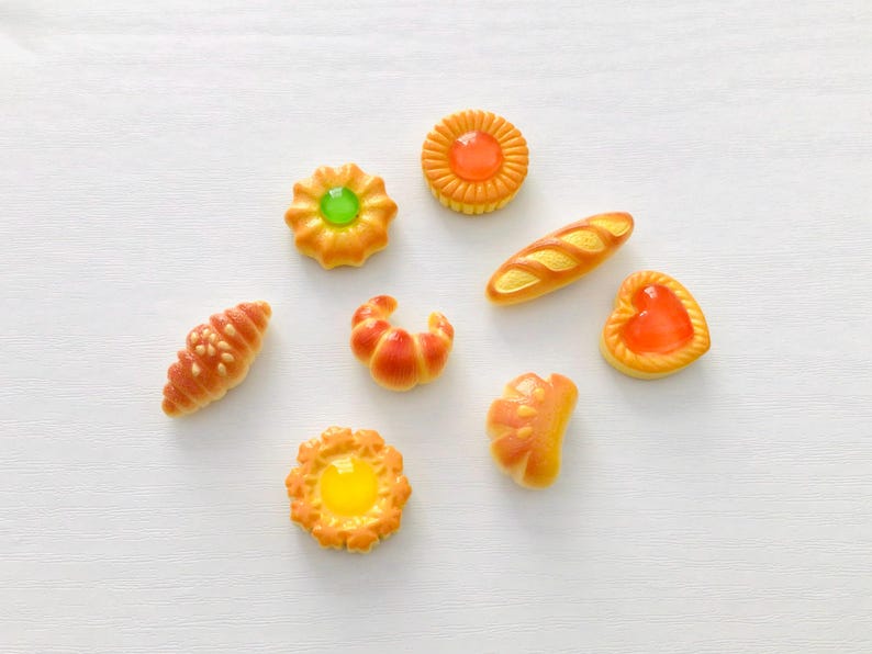 May include: A selection of miniature baked goods, including croissants, bread, and tarts, in shades of orange and yellow. The pastries are arranged on a white surface, showcasing their detailed designs and textures. A delightful assortment of miniature food items.