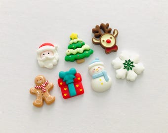 May include: A collection of seven holiday-themed flatback resin charms including a Santa Claus face, a green Christmas tree, a brown reindeer, a gingerbread person with a scarf, a red gift box, a snowman, and a white snowflake with green detail.