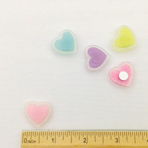 Peut inclure: Plusieurs d&eacute;corations en forme de c&oelig;ur aux couleurs pastel, rose, bleu, violet et jaune. Chaque c&oelig;ur a un int&eacute;rieur scintillant et un contour transparent. Une r&egrave;gle gradu&eacute;e en pouces est visible en bas.