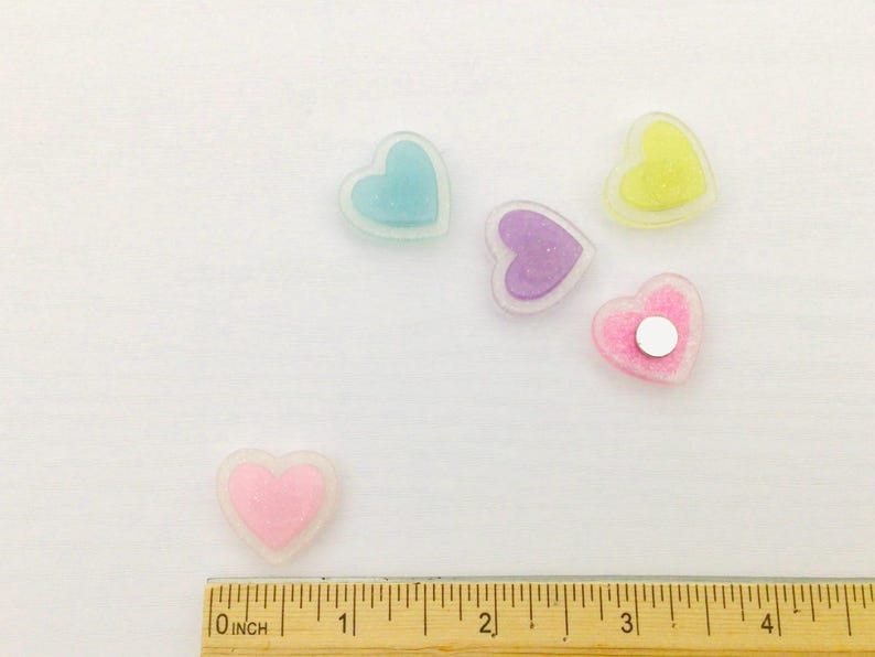 Peut inclure: Plusieurs d&eacute;corations en forme de c&oelig;ur aux couleurs pastel, rose, bleu, violet et jaune. Chaque c&oelig;ur a un int&eacute;rieur scintillant et un contour transparent. Une r&egrave;gle gradu&eacute;e en pouces est visible en bas.