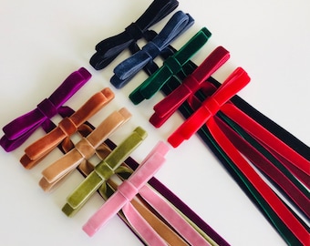 May include: A collection of velvet ribbon bows in various colors including black, navy, dark green, red, purple, brown, beige, olive green, and light pink. Each bow features long, trailing ribbon tails extending downwards from the center knot.