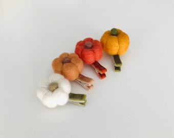 May include: Four felt pumpkin hair clips in shades of orange, yellow, and white. Each clip has a different colored felt pumpkin attached to a metal clip with a velvet ribbon. The pumpkins have a stem detail.