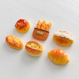 May include: A collection of miniature bread items, including a round bun, baguette, loaf, croissant, and a bread basket with baguettes. The bread pieces are painted in shades of yellow, orange, and brown, with white accents on some. The word "Bread" is visible on the basket.