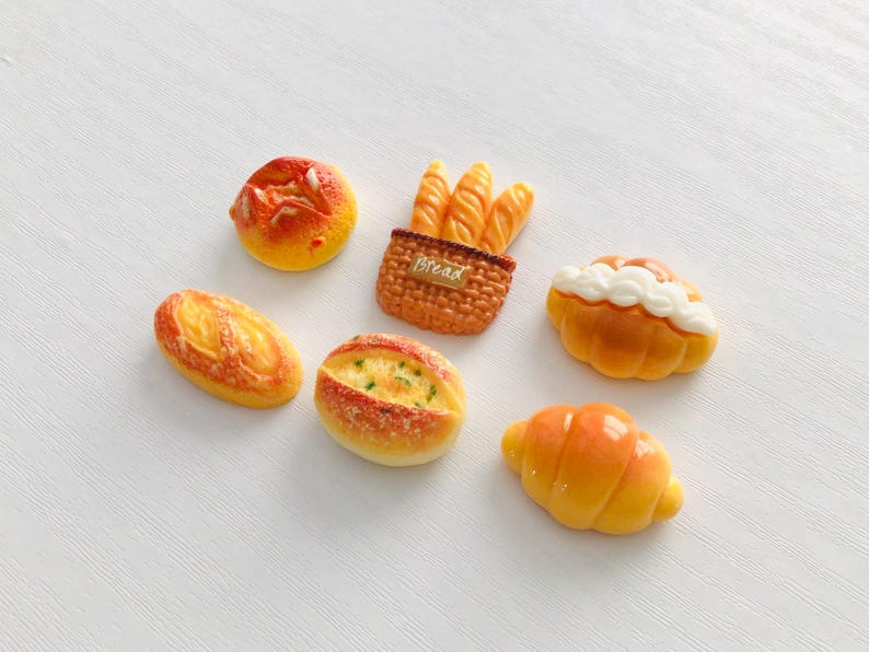 May include: A collection of miniature bread items, including a round bun, baguette, loaf, croissant, and a bread basket with baguettes. The bread pieces are painted in shades of yellow, orange, and brown, with white accents on some. The word "Bread" is visible on the basket.