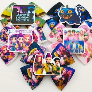 May include: A collection of colorful hair bows featuring various K-Pop and anime-style graphics. The bows display images of cartoon characters, groups, and the text "K-POP DEMON HUNTERS" and "HUNTRIX".