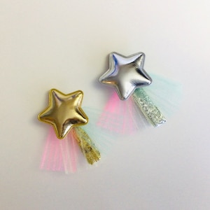 May include: Two gold and silver star-shaped hair clips with tulle tails. The gold star has a pink and gold tulle tail, and the silver star has a light blue and silver tulle tail.