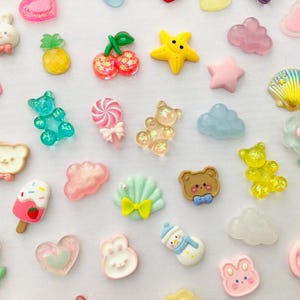 Peut inclure: Une collection d'embellissements en r&eacute;sine color&eacute;e de diff&eacute;rentes formes, dont une &eacute;toile jaune, des c&oelig;urs roses et un bonhomme de neige bleu. D'autres formes incluent un ours, un lapin et un coquillage.