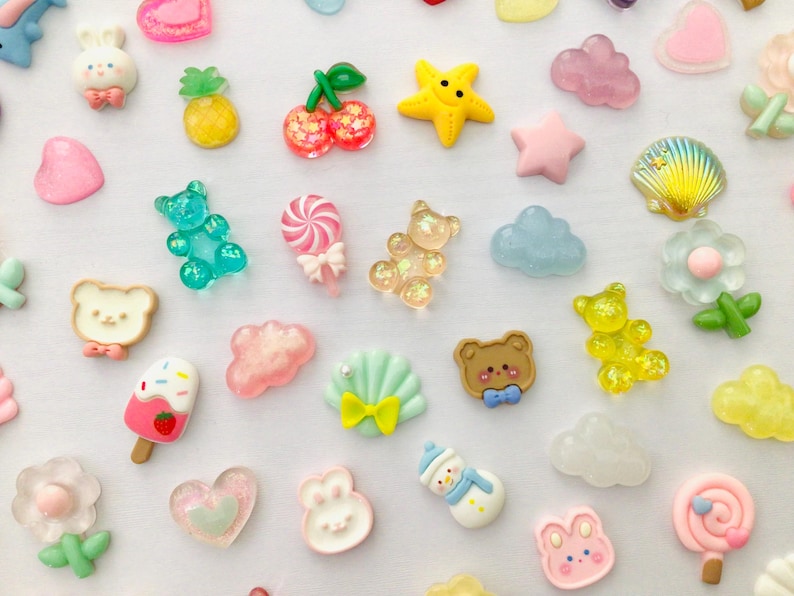 Peut inclure: Une collection d'embellissements en r&eacute;sine color&eacute;e de diff&eacute;rentes formes, dont une &eacute;toile jaune, des c&oelig;urs roses et un bonhomme de neige bleu. D'autres formes incluent un ours, un lapin et un coquillage.