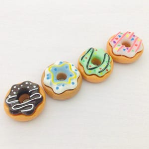 May include: Four miniature donuts with colourful icing. One donut has chocolate icing with white drizzle and white sprinkles. Another has blue icing with yellow sprinkles. The third has green icing with green drizzle. The last has pink icing with rainbow sprinkles.