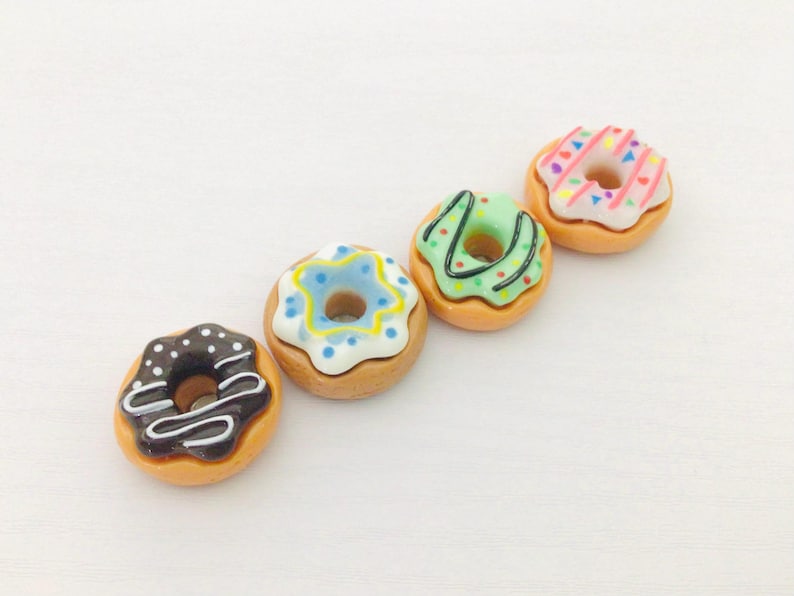 May include: Four miniature donuts with colourful icing. One donut has chocolate icing with white drizzle and white sprinkles. Another has blue icing with yellow sprinkles. The third has green icing with green drizzle. The last has pink icing with rainbow sprinkles.