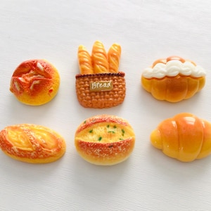 May include: A collection of miniature bread-themed decorative items. The assortment includes a round loaf, a baguette basket with the word "Bread", a pastry with white frosting, a long oval loaf, a round loaf with green specks, and a croissant.