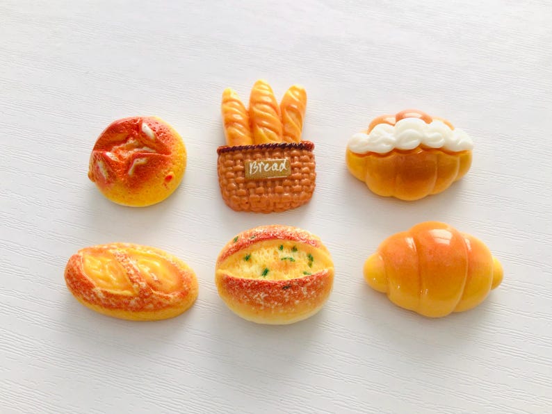 May include: A collection of miniature bread-themed decorative items. The assortment includes a round loaf, a baguette basket with the word "Bread", a pastry with white frosting, a long oval loaf, a round loaf with green specks, and a croissant.