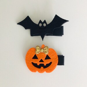 May include: Two Halloween hair clips, one shaped like a black bat and the other shaped like an orange pumpkin with a gold glitter bow.