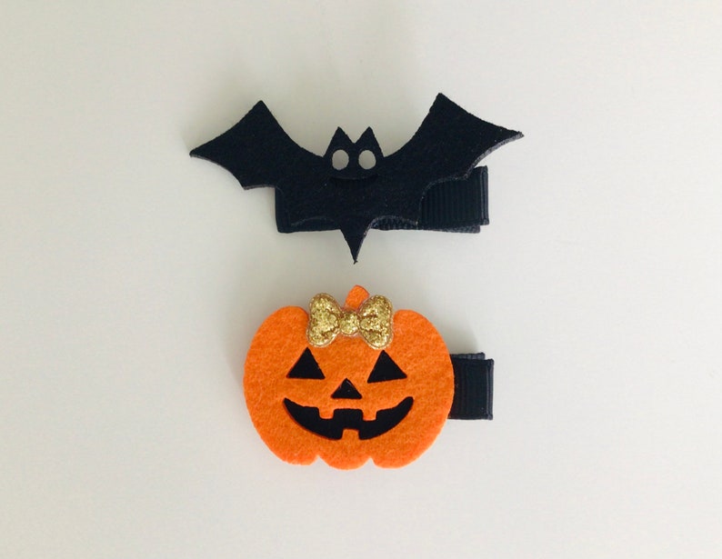 May include: Two Halloween hair clips, one shaped like a black bat and the other shaped like an orange pumpkin with a gold glitter bow.