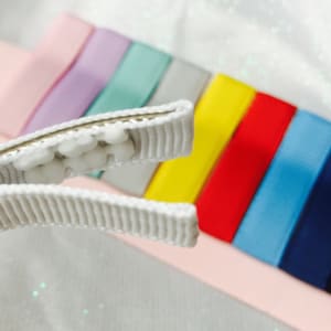 May include: A white alligator clip with a row of white beads. The clip is open and in focus. The background is blurred and shows a rainbow of colored ribbons.