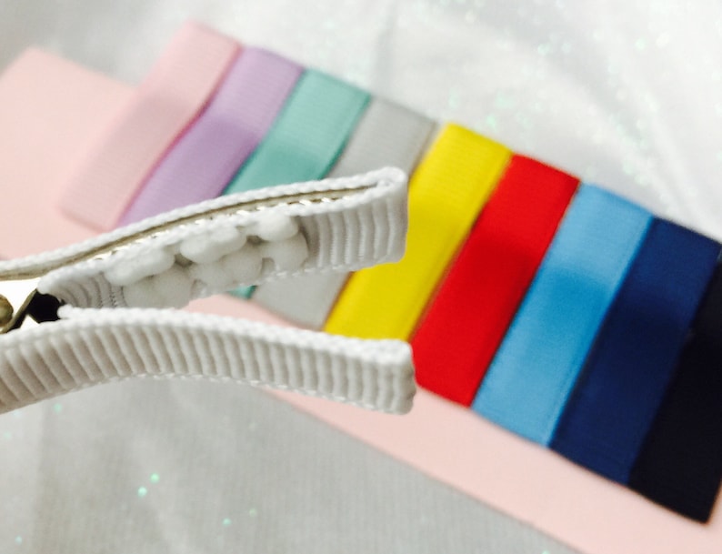 May include: A white alligator clip with a row of white beads. The clip is open and in focus. The background is blurred and shows a rainbow of colored ribbons.