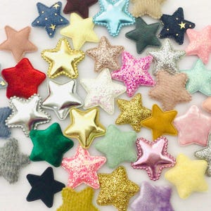 May include: A collection of assorted star-shaped embellishments in various colors and textures. The stars include denim, glitter, velvet, and metallic finishes. Colors range from pink, gold, and silver to blue, green, and yellow. These decorative stars are ideal for crafts.